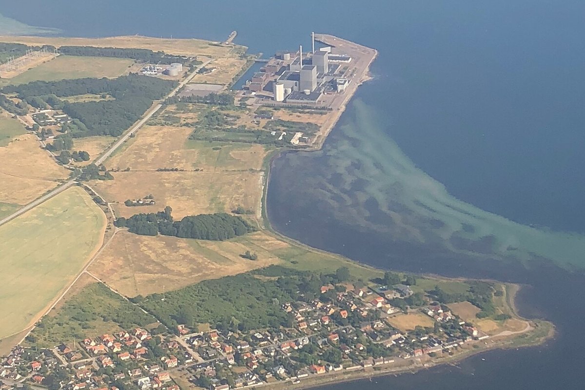 Luftfoto af et atomkraftværk med store grå bygninger og skorstene på en landtange ud i Øresund, med marker og et villakvarter i forgrunden