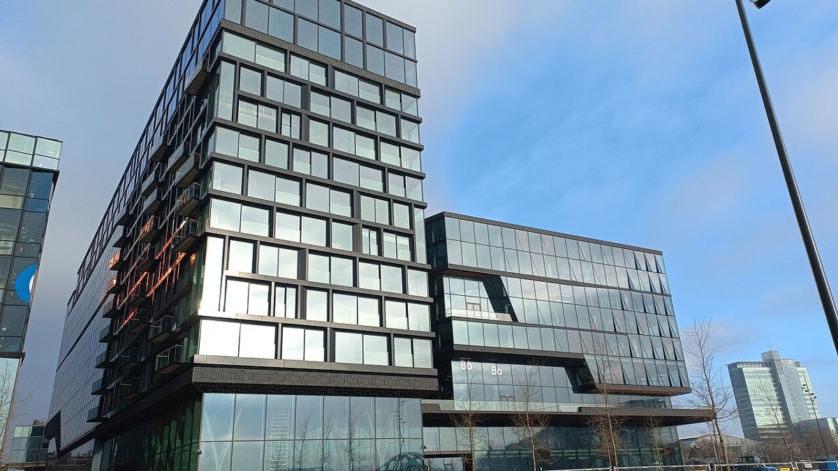 Booking.coms hovedkontor, en høj bygning af glas og mørke rammer, på Oosterdokseiland i Amsterdam under blå himmel