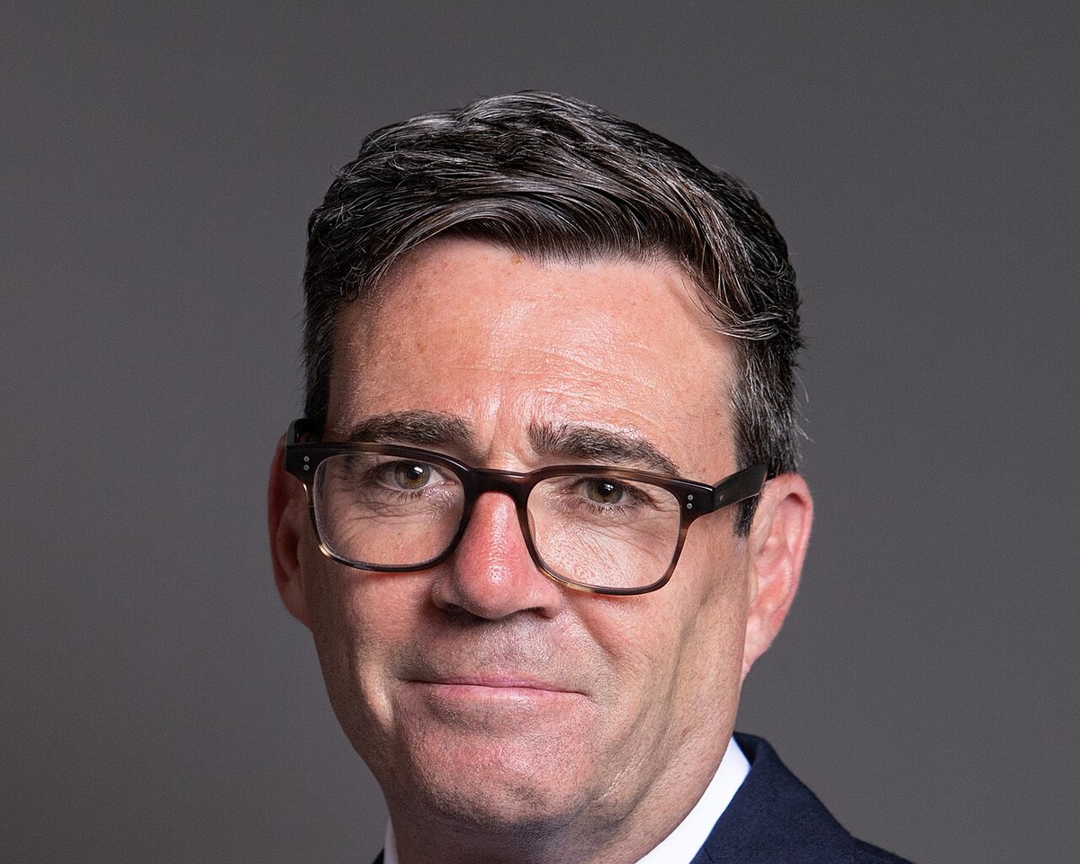 Portræt af Andy Burnham med mørkt hår og briller med brunt stel foran en grå baggrund