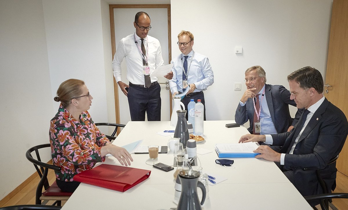 Mette Frederiksen og Mark Rutte sidder ved et hvidt mødebord med papirer, mens to rådgivere står bag dem og en tredje sidder ved bordet