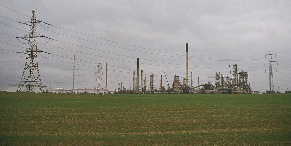 Raffinaderiet i Kalundborg med skorstene og procesanlæg set fra en grøn mark under en grå himmel med højspændingsmaster i forgrunden