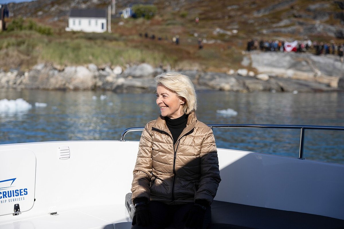 Ursula von der Leyen i brun dunjakke står på dækket af en båd med vand, klipper og is bag sig